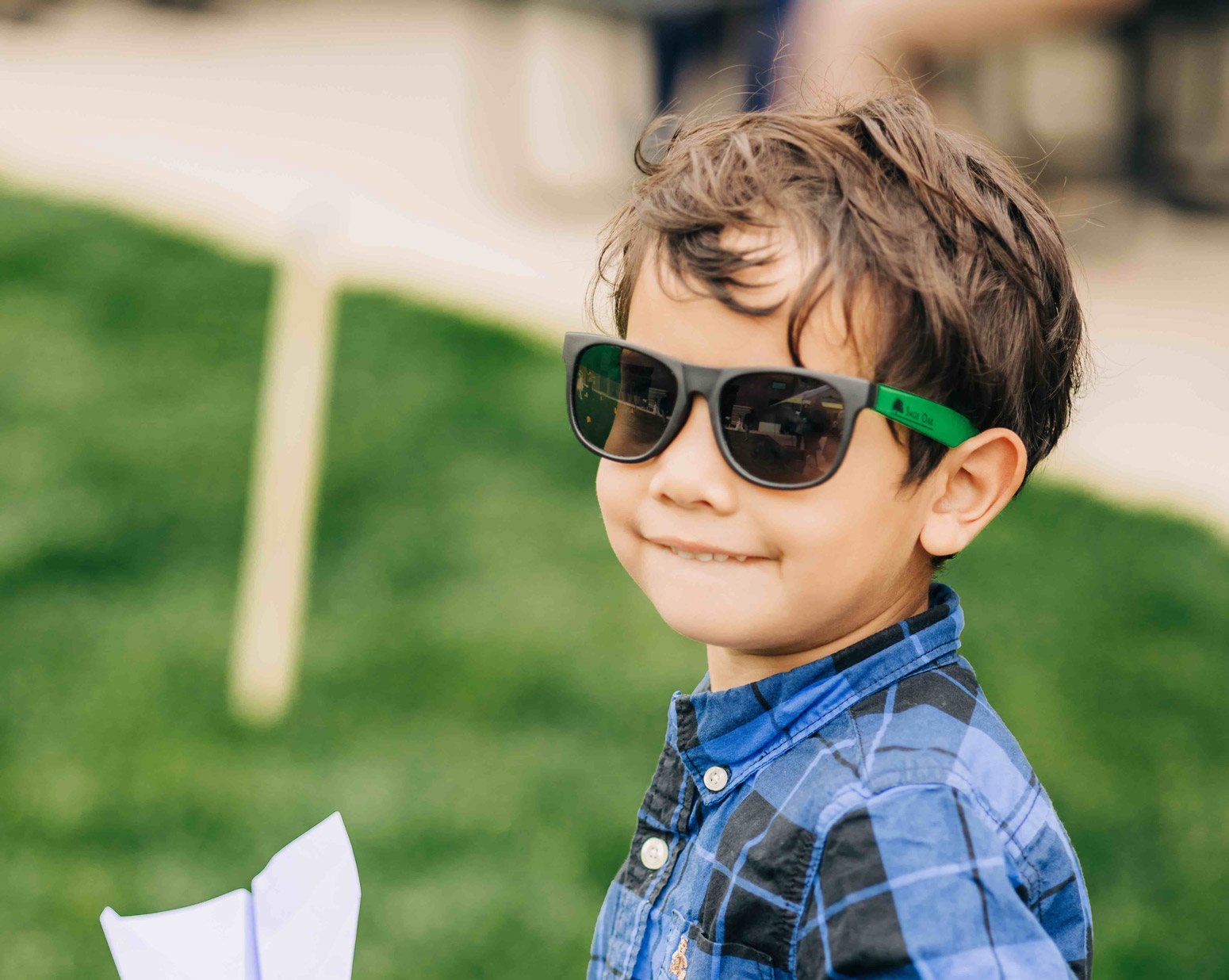 young boy in sunglasses smiling as he holds a paper airplane